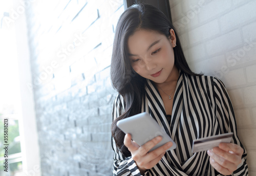 Asian young woman using smartphone and gray credit card to online shopping, transfer money, financial, internet banking. Background sunlight and copy space. © Pravinrus