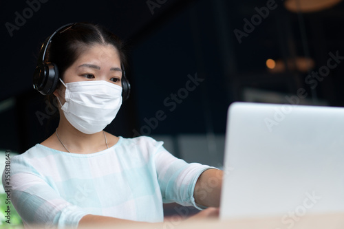 young asian woman wearing face mask and headphone and using computer to work from home during covid-19 or coronavirus outbreak. social distancing and new normal lifestyle comcept