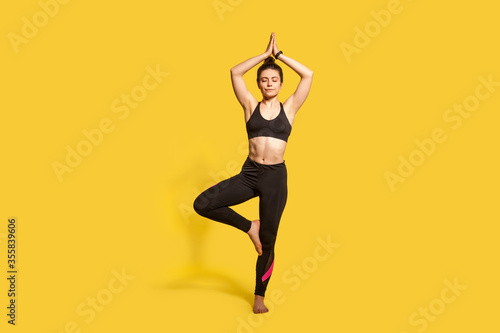 Tree pose. Athletic concentrated woman in tight sportswear practicing yoga, meditating with closed eyes, doing Vrksasana exercise on one leg, hands up. full length studio shot, sport workouts isolated