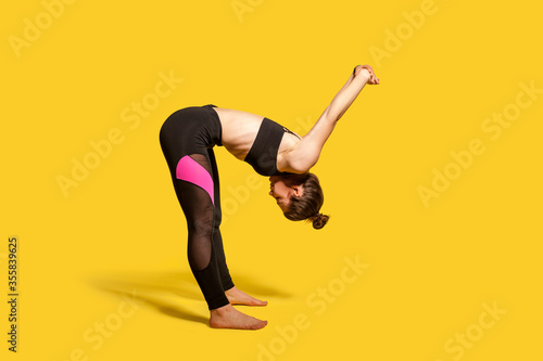 Athletic girl with hair bun in tight sportswear doing sport, bending and stretching hands backwards, warming up training muscles for flexibility. full length studio shot, isolated on yellow background