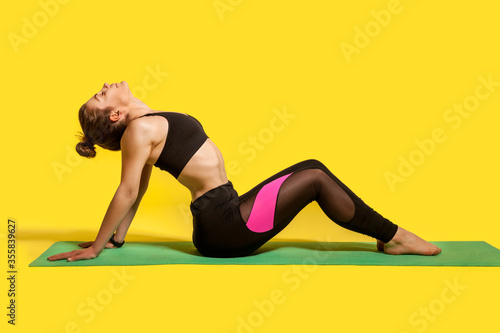 Fitness woman with hair bun in tight sportswear sitting on gym mat doing sport, bending backwards stretching spinal muscles, warming up with flexibility exercises. studio shot, isolated on yellow