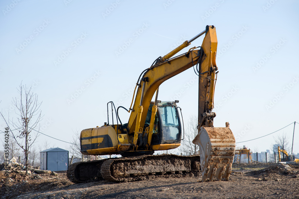 Excavator at building under construction. Backhoe digs the ground for the foundation and for laying sewer pipes. Renovation program. Buildings industry background Excavator at building under construction. Backhoe digs the ground for the foundation and for laying sewer pipes. Renovation program. Buildings industry background