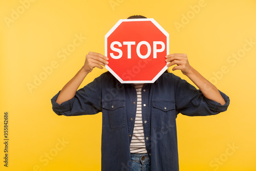 Attention, prohibited way! Unknown person in denim shirt hiding face behind Stop symbol, red traffic sign warning of restricted access, banned service. indoor studio shot isolated on yellow background
