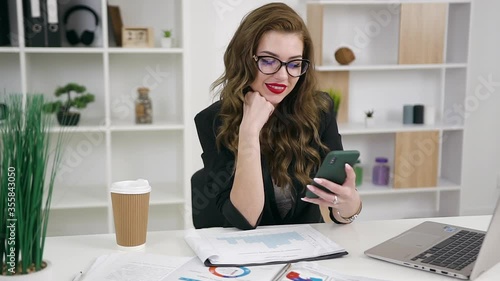 Portrait of attractive confident positive young business lady in glasses dressed in trendy jacket with brightly red lipstick using her phone in office room photo