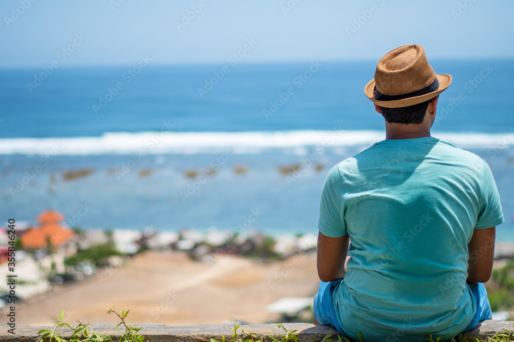 A tourist sitting on a cliff and enjoying a hot summer day wearing colourful dress and round hat beside a sea beach in Bali