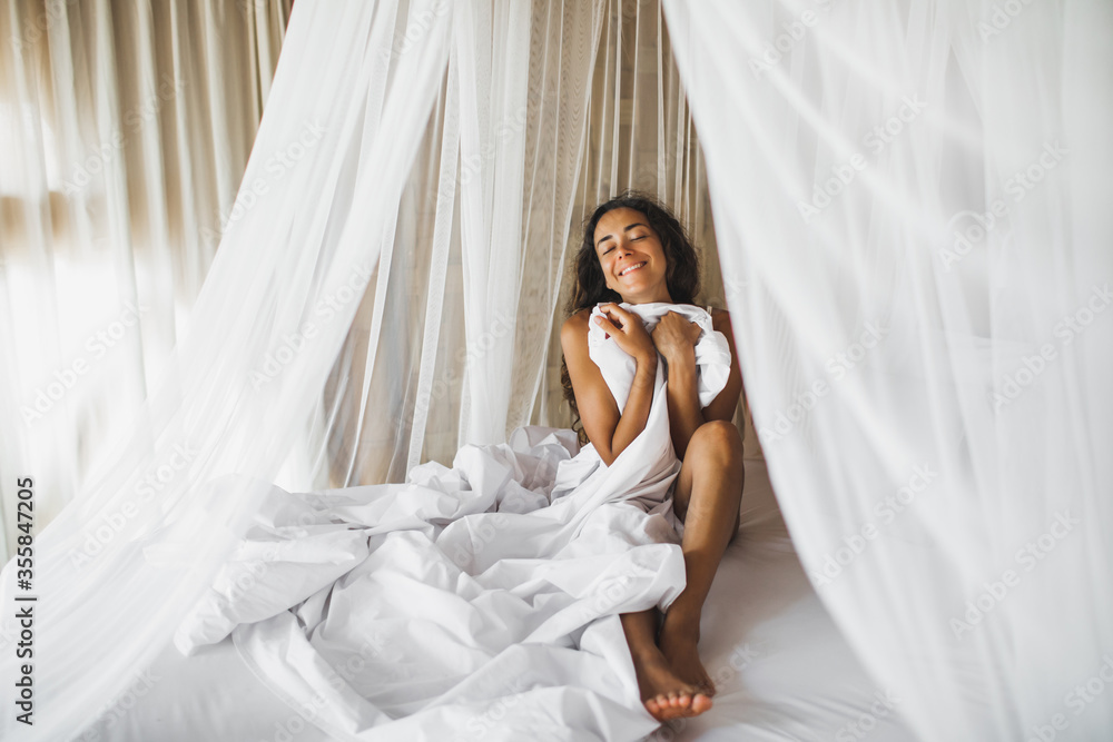 Beautiful tanned young woman awakening in white bed. Happy wake up and start new day. Leisure and rest. Wellbeing and carefree concept. Long curly hair. Stay home. Beautiful tanned young woman awakening in white bed. Happy wake up and start new day. Leisure and rest. Wellbeing and carefree concept. Long curly hair. Stay home.