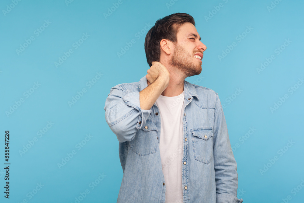 Portrait of exhausted young man in denim shirt touching neck and grimacing from intense pain, massaging to relieve muscle tension, hurting shoulders. indoor studio shot isolated on blue background Portrait of exhausted young man in denim shirt touching neck and grimacing from intense pain, massaging to relieve muscle tension, hurting shoulders. indoor studio shot isolated on blue background