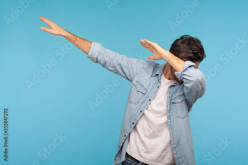 Dab dance. Portrait of man in denim shirt making dabbing movement, famous internet meme of success victory, expressing happiness and following trends. indoor studio shot isolated on blue background photo