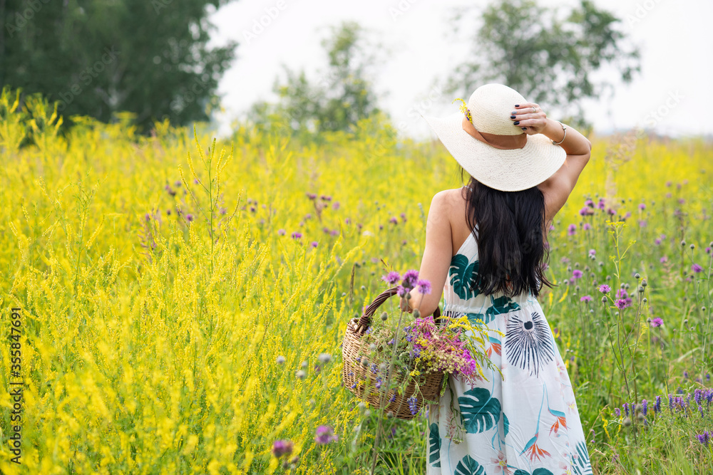 A girl with a full basket of flowers in a light long summer dress stands in the middle of a yellow flower field. Bright summer colors. The enjoyment of nature