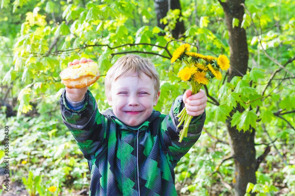 The boy hands yellow flowers dandelions and a delicious doughnut to his mother as a gift. The boy hands yellow flowers dandelions and a delicious doughnut to his mother as a gift.