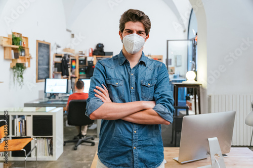 Portrait of businessman in office with arms crossed near work station with medical mask in protection from contamination by Coronavirus, Covid-19 - Millennial man at working for his start up company photo