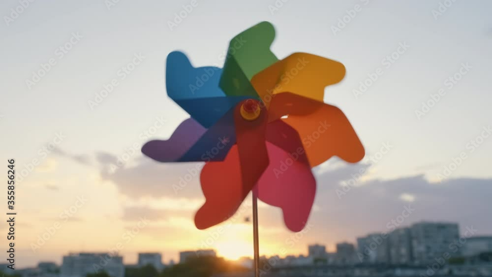 Pinwheel rotating colored plastic with blowing wind against a blue sky, white clouds, silhouettes of city buildings on sunset slow motion. Symbol of freedom and life. Toy spinner. Bright disk of sun Pinwheel rotating colored plastic with blowing wind against a blue sky, white clouds, silhouettes of city buildings on sunset slow motion. Symbol of freedom and life. Toy spinner. Bright disk of sun
