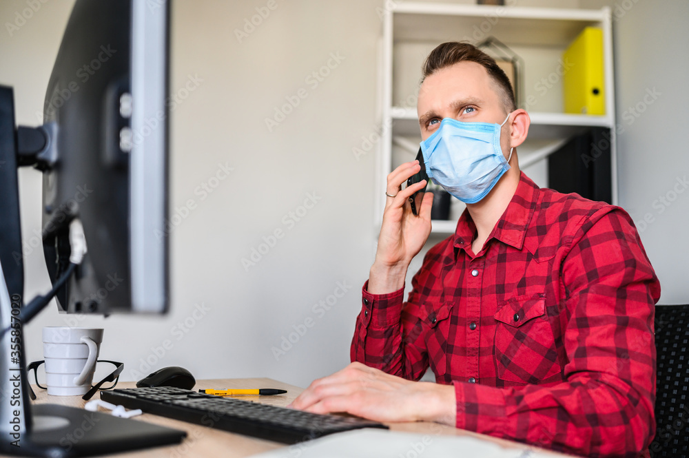 Responsible employee talks on the phone while sitting at the office desk, he wears a protective medical mask. A young man works in the office while quarantine with the PC