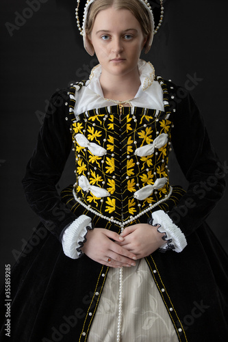 A young woman in black velvet Elizabethan costume standing against a dark studio backdrop