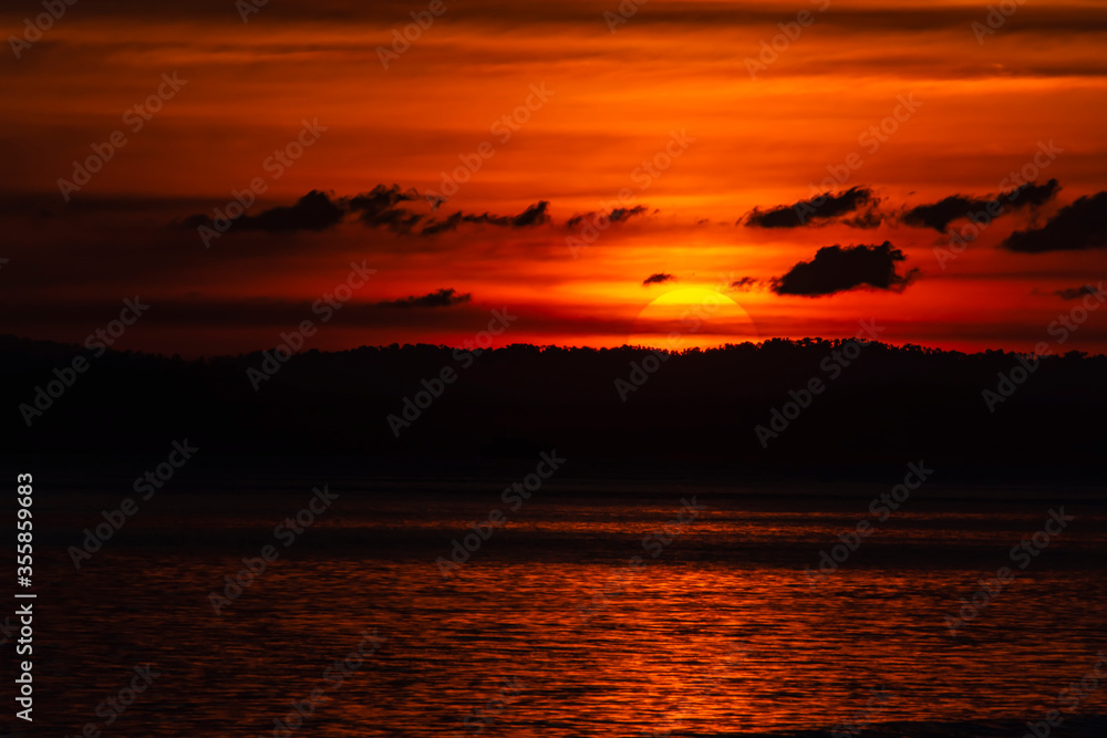 Magical & dramatic scenic landscape shot of sun setting down, only half visible, behind a silhouette of mountains & clouds and the sunshine sparkling in the water. A stunning orange & black scenery.