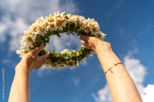 A wreath of white daisies in a woman's hand against a background of blue sky and white clouds