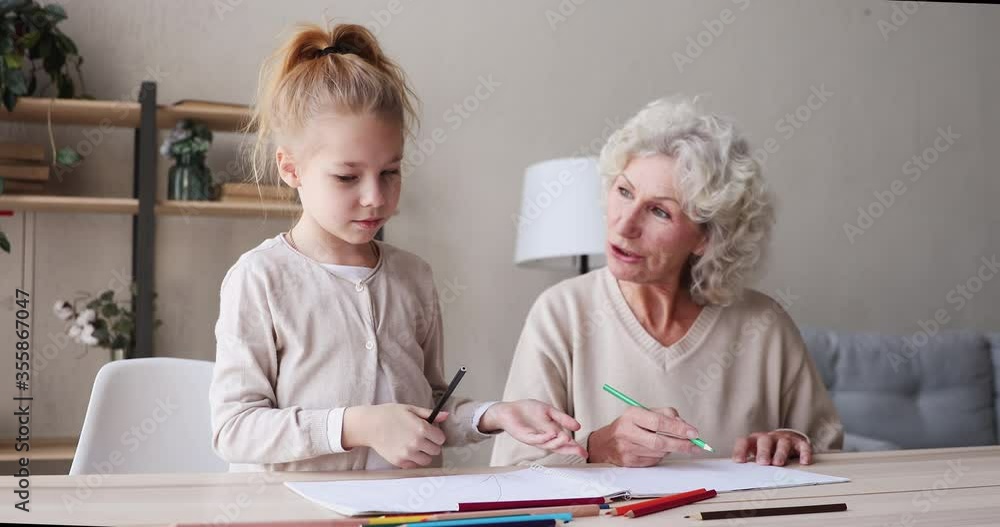 Happy 7 years old adorable small child girl drawing in paper album with colorful pencils together with affectionate caring middle aged mature grandma in living room, weekend childcare daycare concept. Happy 7 years old adorable small child girl drawing in paper album with colorful pencils together with affectionate caring middle aged mature grandma in living room, weekend childcare daycare concept.