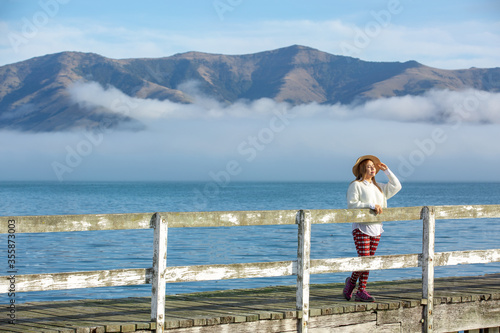 Beautyful asian woman resting is looks at the sea bay with houses and boats on a sunny day at Childrens bay, Akaroa, Canterbury, New Zealand. Relaxing young woman at harbor. © tonklafoto