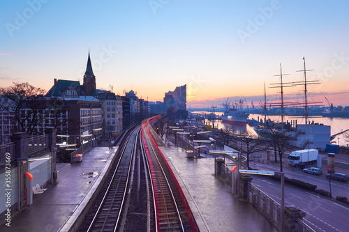 Landungsbrucken station in Hamburg, Germany on a sunrise, early in the morning, with tracer of rear lights of passing train. Elbe riverside, downtown skyline at dawn, on sunrise.