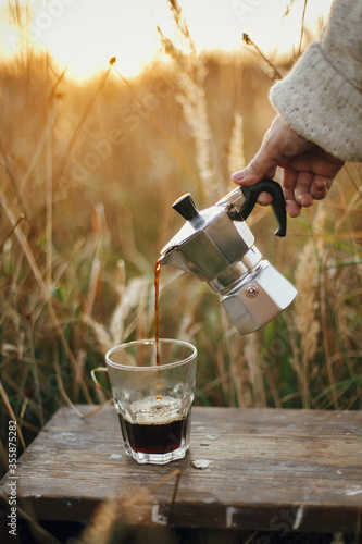Hipster pouring fresh hot coffee from geyser coffee maker into glass cup in sunny warm light in rural countryside herbs. Atmospheric tranquil moment. Alternative coffee brewing in travel