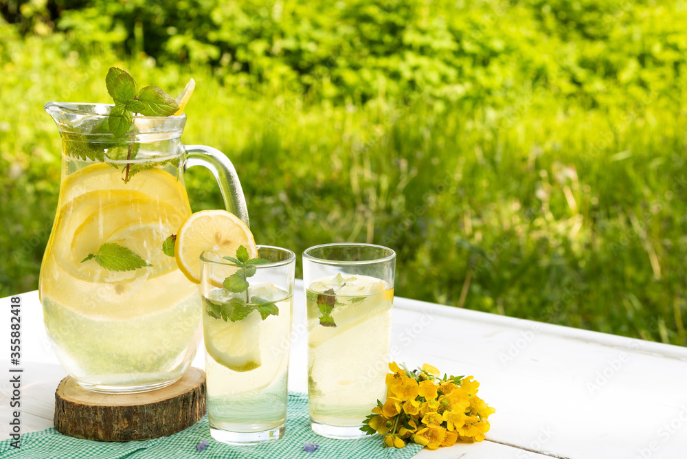 Two glasses and a pitcher of lemonade on the street against the background of greenery on a hot summer day. Two glasses and a pitcher of lemonade on the street against the background of greenery on a hot summer day.