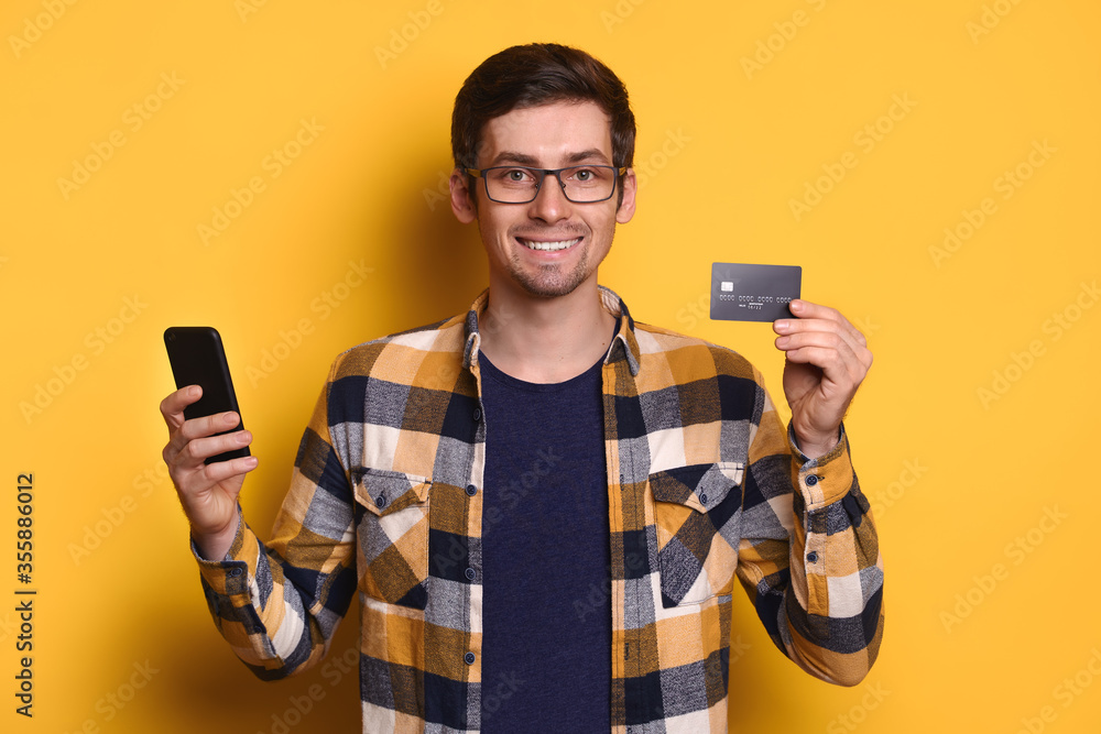 Young caucasian man in glasses using mobile phone and credit card for online shopping, e-commerce, online banking, smiling isolated over yellow studio background. Technology and reaction concept Young caucasian man in glasses using mobile phone and credit card for online shopping, e-commerce, online banking, smiling isolated over yellow studio background. Technology and reaction concept