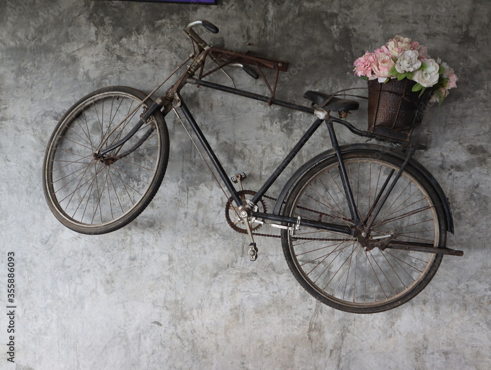 The old black bicycle with a flower basket at the end is hung on a concrete wall to decorate the place to look beautiful. The old black bicycle with a flower basket at the end is hung on a concrete wall to decorate the place to look beautiful.