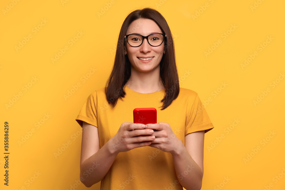 Smiling young woman with brown hair in eyeglasses types text message on smartphone, enjoys online communication, writes feedback, wears orange t-shirt, isolated on yellow wall. Technology concept