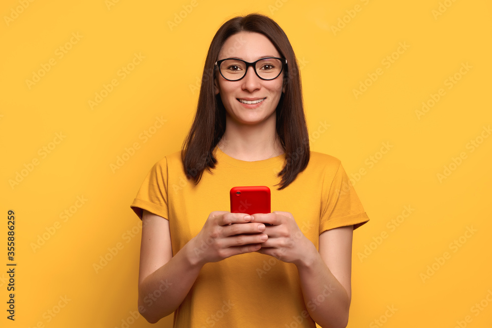 Smiling young woman with brown hair in eyeglasses types text message on smartphone, enjoys online communication, writes feedback, wears orange t-shirt, isolated on yellow wall. Technology concept Smiling young woman with brown hair in eyeglasses types text message on smartphone, enjoys online communication, writes feedback, wears orange t-shirt, isolated on yellow wall. Technology concept
