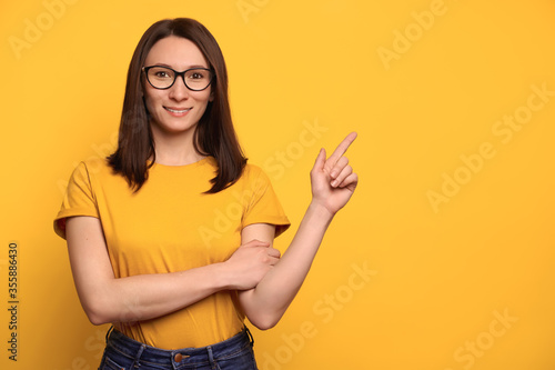 Beautiful smiling girl in eyeglasses pointing with index finger at copy space for advertisement, isolated on yellow background. Pretty girl showing recommendation, advising product. Promotion concept