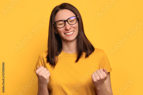 Young brunette woman in glasses and yellow t-shirt posing isolated on yellow background, doing winner yes gesture, studio portrait. People sincere emotions and winner concept. Mock up copy space