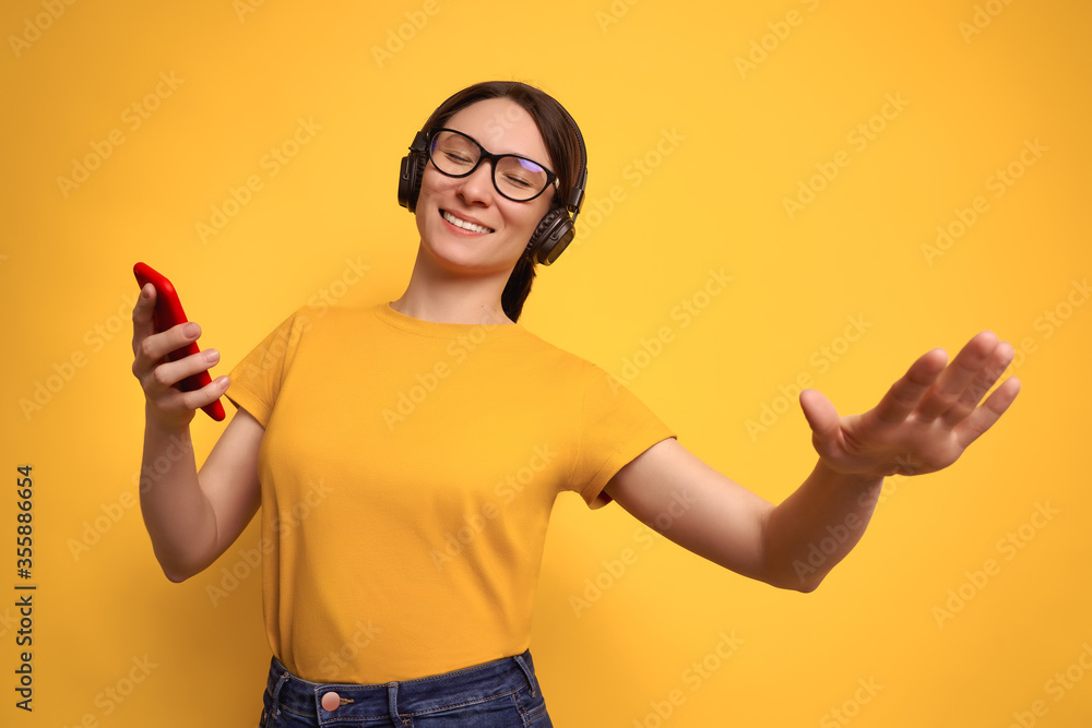 Studio shot of beautiful brunette woman in yellow t-shirt and wireless headphones listens to loud music and dancing on yellow background. Joyful girl enjoy favorite playlist with online smartphone app Studio shot of beautiful brunette woman in yellow t-shirt and wireless headphones listens to loud music and dancing on yellow background. Joyful girl enjoy favorite playlist with online smartphone app