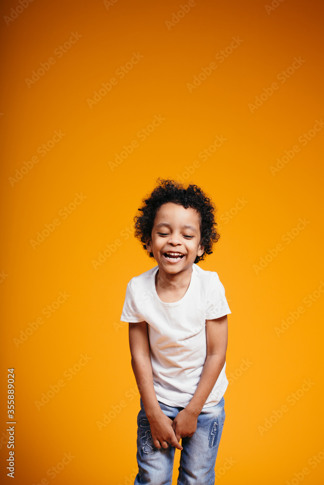 Curly-haired Ethiopian boy in a white T-shirt straightens jeans, laughs and closed his eyes on an orange background in the studio Curly-haired Ethiopian boy in a white T-shirt straightens jeans, laughs and closed his eyes on an orange background in the studio