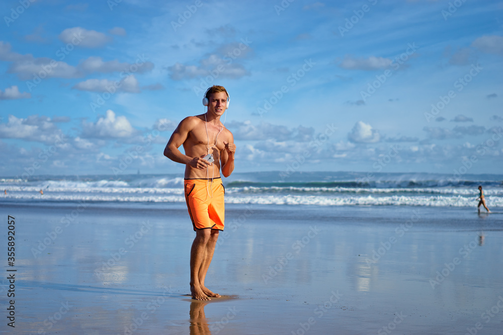 Enjoying music. Sports lifestyle. Happy young man in headphones walking on the sea shore.