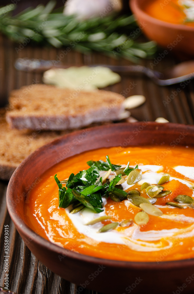 Homemade rustic pumpkin soup with seeds in clay dish on wooden table with bread spices and greens aside Homemade rustic pumpkin soup with seeds in clay dish on wooden table with bread spices and greens aside