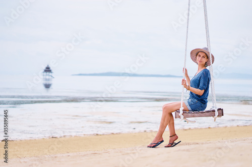Vacation concept. Happy young woman in hat sitting on swing enjoying sea view.