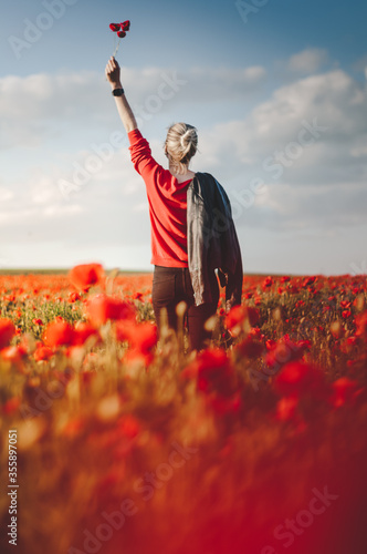 Beautiful young blonde girl victory holding a poppies bouquet in poppy field at sunset in leather jacket from behind © Jan