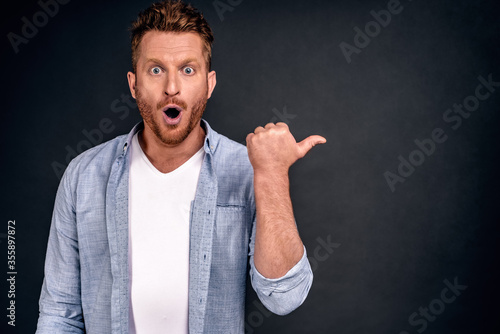 Horizontal shot of amazed young man has thick red beard, dressed in fashionable clothes, indicates with thumb aside, shocked to notice something, copy space for your promotional text or hearder photo