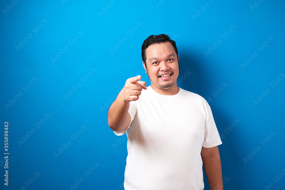 Portrait of young Asian man in white t-shirt smiling and pointing forward, looking at camera. Choosing someone concept with selective focus, against blue background Portrait of young Asian man in white t-shirt smiling and pointing forward, looking at camera. Choosing someone concept with selective focus, against blue background