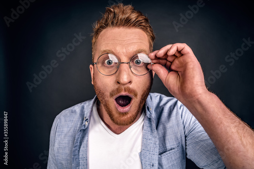 Close up portrait of stunned bearded young redhead guy drops jaw, has bugged blue eyes, sees something unbelievable and surprising, has eyewear, isolated on grey background. People, emotions concept