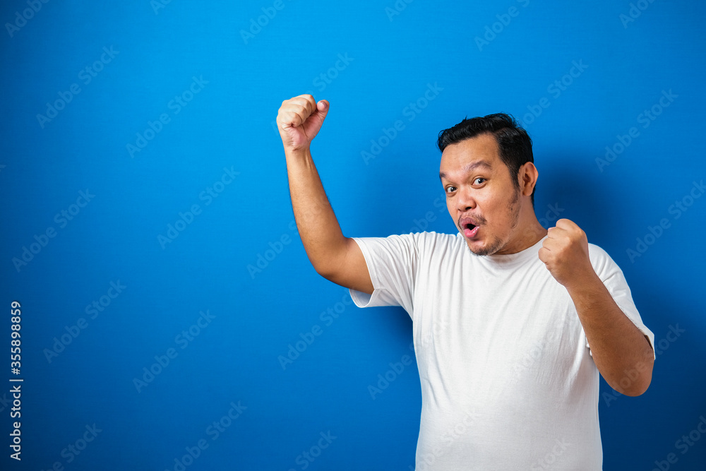 Portrait of a funny fat Asian man in white t-shirt smiling and dancing happily, joyful expressing celebrating good news victory winning success gesture against blue background Portrait of a funny fat Asian man in white t-shirt smiling and dancing happily, joyful expressing celebrating good news victory winning success gesture against blue background