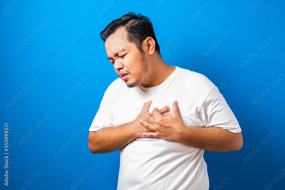 Portrait of good looking young Asian man in white t-shirt having pain on his chest, against blue background.