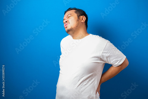 Portrait of good looking young Asian man in white t-shirt having pain on his backs, against blue background.