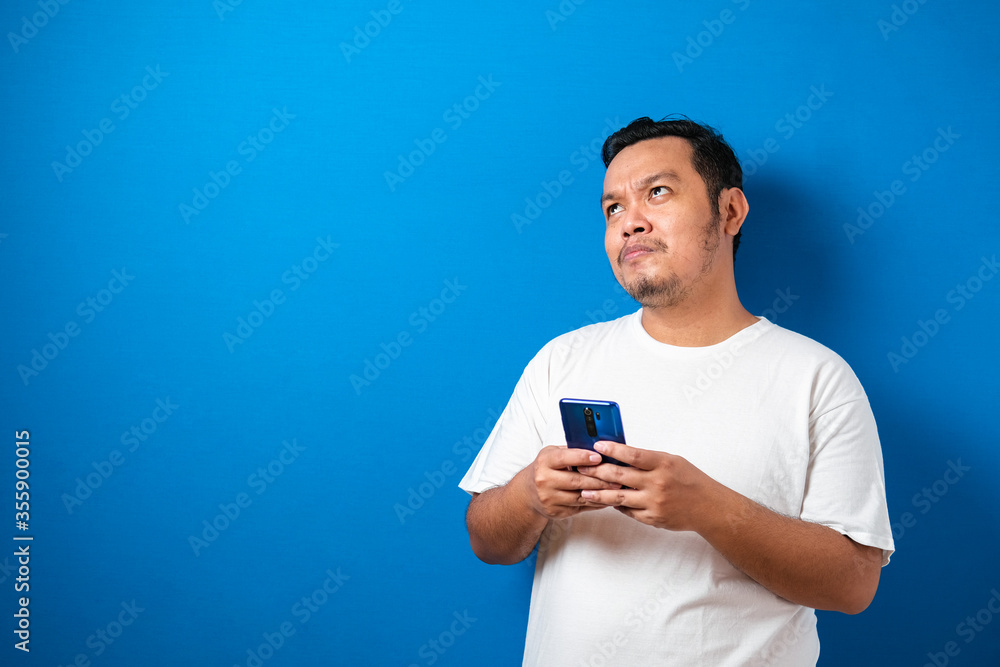 Fat Asian man in white t-shirt, thinking something while chat messaging on his smart phone, half body portrait over blue background