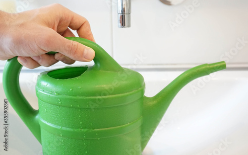 A hand grips a filled watering can in a bathroom sink to water indoor plants.