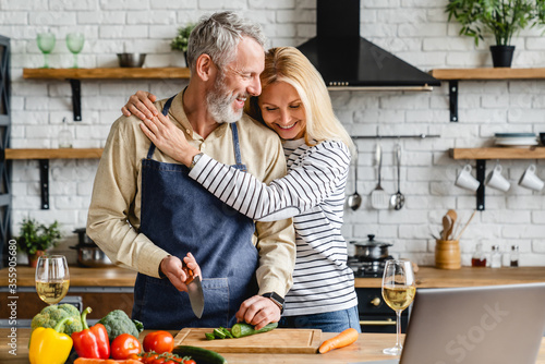 Beautiful mature couple hugging while cooking vegetable salad at kitchen table photo