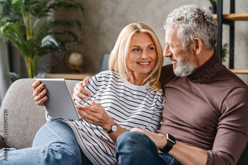 Happy middle aged couple using digital tablet on sofa at home while looking on each other