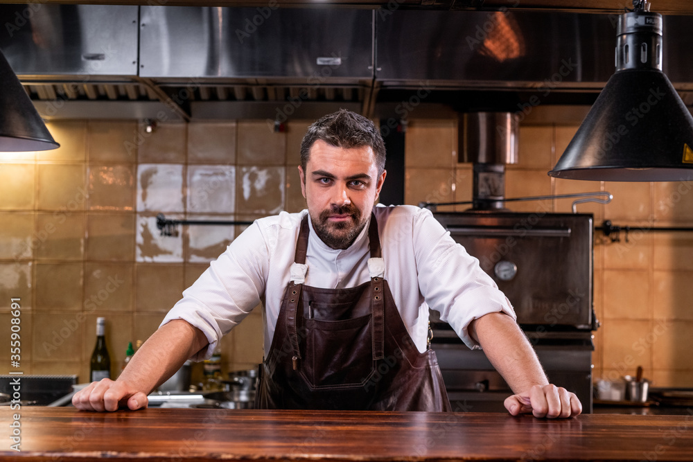 Portrait of confident handsome bearded chef in leather apron standing at wooden counter in open kitchen Portrait of confident handsome bearded chef in leather apron standing at wooden counter in open kitchen