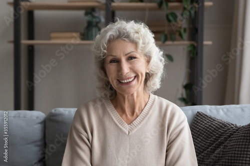 Headshot portrait of smiling middle-aged 60s woman relax on sofa at home, enjoy calm weekend indoors, happy mature 50s grandmother sit rest on couch in living room, feel positive optimistic