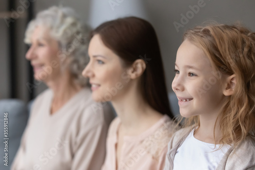 Profile view of happy three generations of women look in distance dreaming visualizing together  hopeful smiling little girl forefront with mom and mature grandmother imagine bright family future